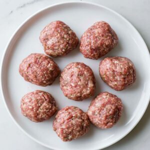 A white plate holding uncooked meatballs, evenly spaced, showing the texture of the ground meat mixture.