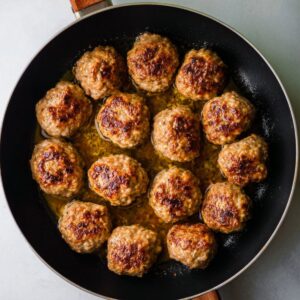 A frying pan filled with golden-brown cooked meatballs, arranged in a circular pattern, sitting in a thin layer of juices.