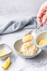 Creamy lemon sauce with fresh lemon wedge and ingredients for healthy recipes on a white marble surface.
