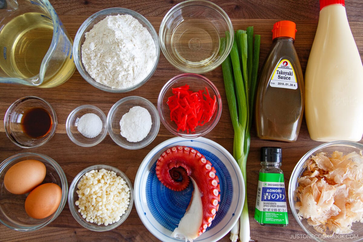 Ingredients for takoyaki are arranged on a wooden surface, including flour, eggs, dashi, octopus, green onions, pickled ginger, bonito flakes, tempura scraps, baking powder, sauces, and seasonings in bowls and bottles.