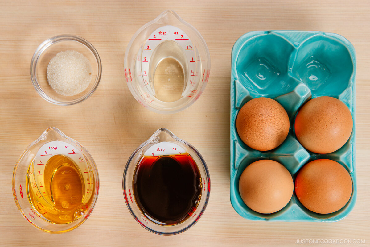 Top-down view of four brown eggs in a blue carton, perfect for making ramen eggs, and four clear containers holding sugar, water, oil, soy sauce, and broth—all neatly arranged on a wooden surface.