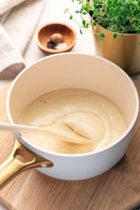A saucepan filled with creamy bechamel sauce is being stirred with a wooden spoon, resting on a wooden board beside a potted herb and a small bowl of spices.