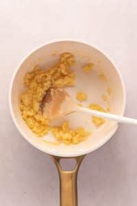 A white saucepan with a gold handle contains a thick, yellow roux being stirred with a wooden spoon-the perfect base for a creamy bechamel sauce-on a light surface.