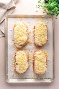 Four slices of bread topped with ham, white sauce, and grated cheese are arranged on a parchment-lined baking sheet, ready to be baked into classic croque monsieur sandwiches.