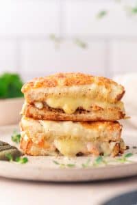 A close-up of a stacked grilled cheese sandwich inspired by croque monsieur, with melted cheese oozing out, placed on a light-colored surface with herbs and pickles nearby.