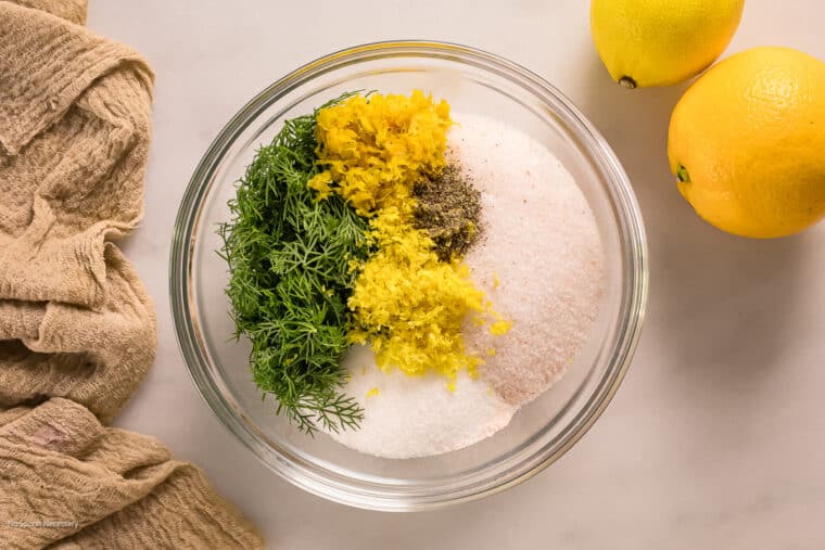 Overhead photo of sugar, salt, fresh dill, lemon zest, orange zest, and black pepper in a medium bowl.