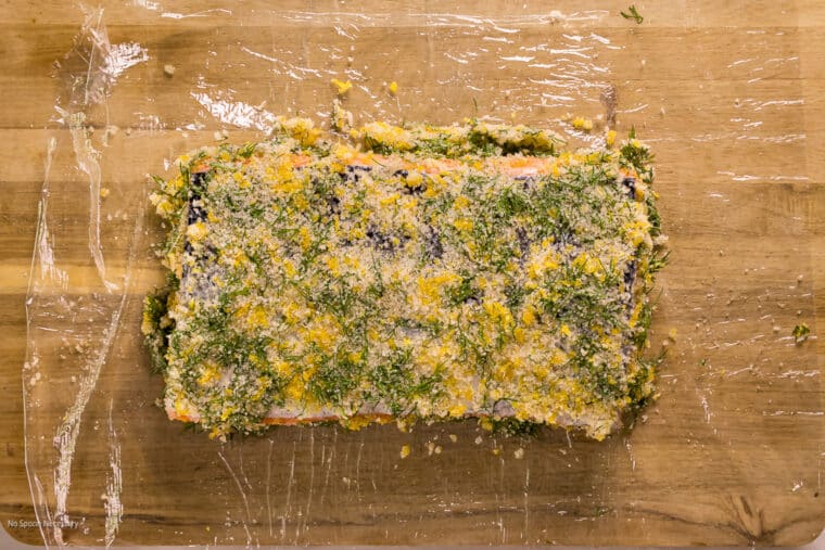 Overhead photo of a slab of raw salmon coated in a salt, sugar, and dill cure on a piece of plastic wrap.