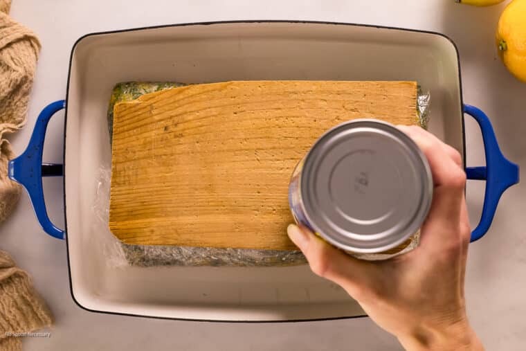 Overhead action photo of a person putting cans on a plastic wrapped cure-coated raw salmon to weigh it down in a pan.