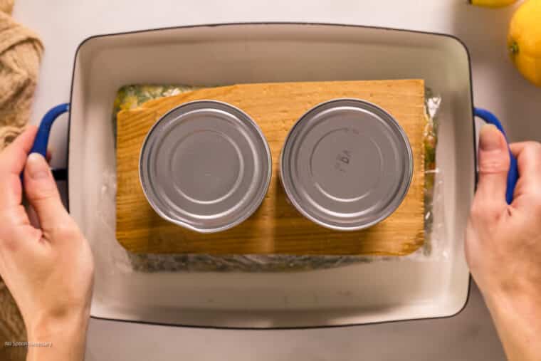 Overhead photo of two hands moving a weighed down piece of curing salmon in a pan from the counter to the fridge.
