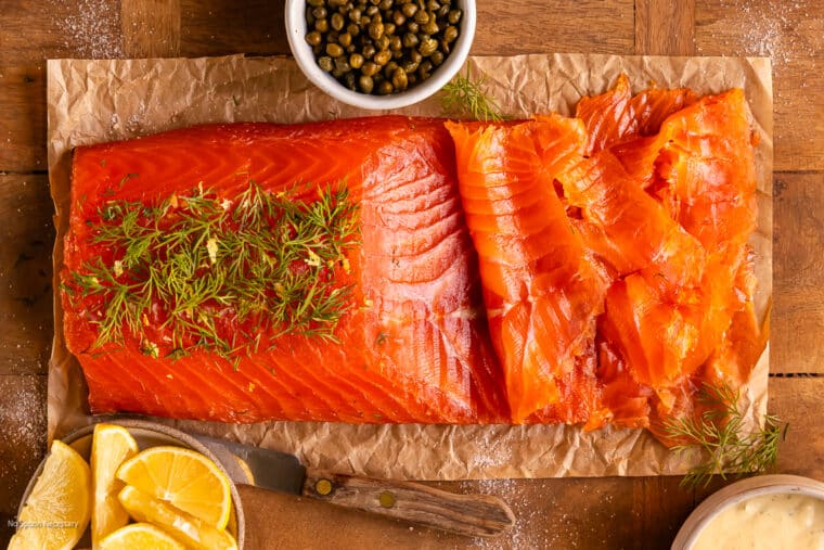 Overhead photo of a partially sliced side of gravlax on a wood board with capers, lemon wedges, and dill.