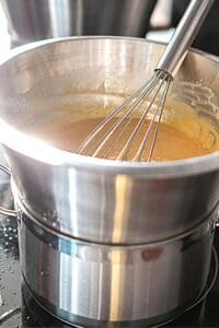 Zabaione recipe preparation: A whisk stirs a creamy, light brown custard in a double boiler. The metal bowl sits atop a pot, showcasing the cooking process for this delicious dessert.